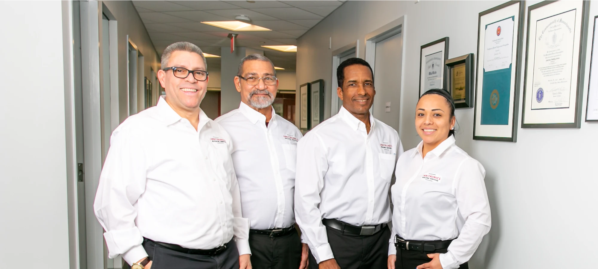 Four Janitronics employees smiling in a hallway with framed certifications on the wall