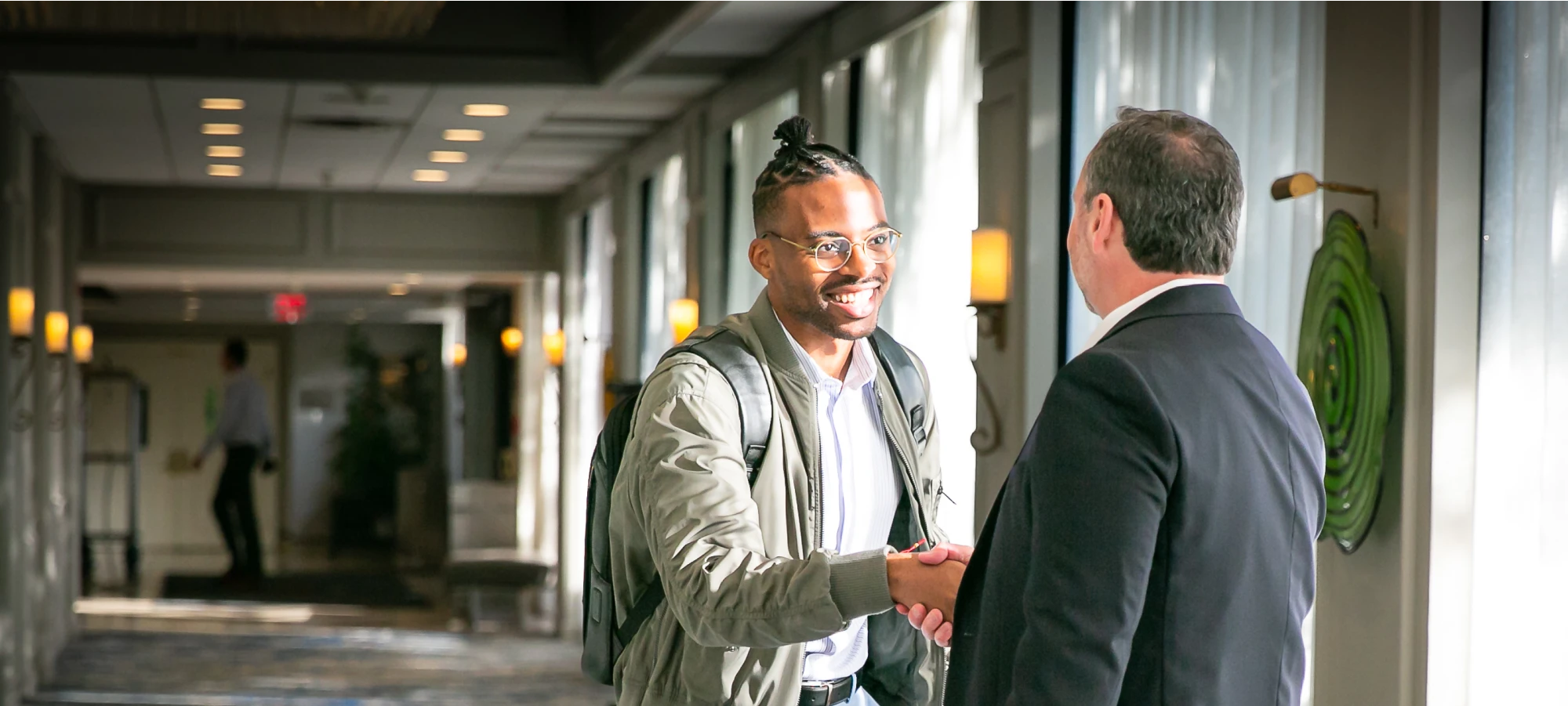 Smiling man with a backpack shakes hands with a business professional in a hallway