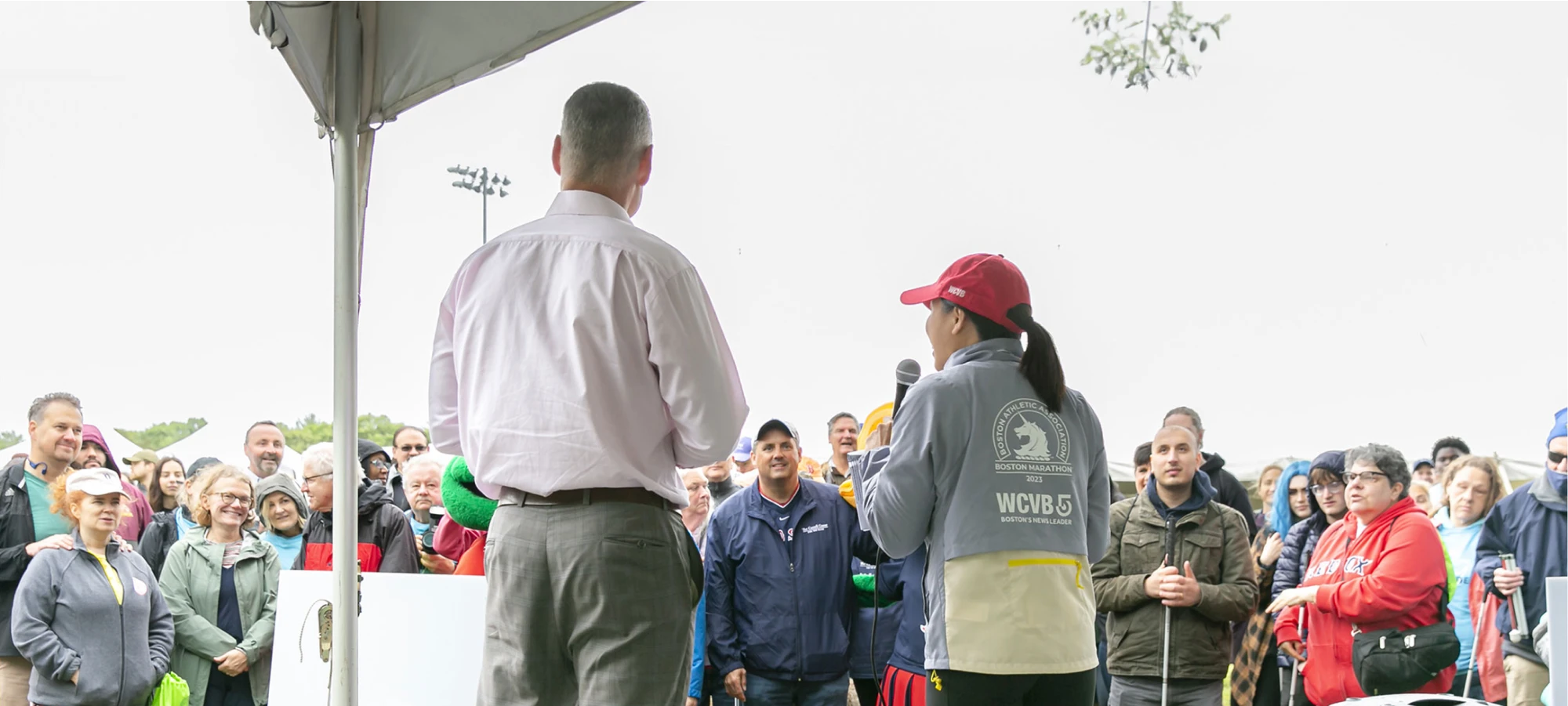 Two speakers address a large outdoor crowd under a tent at a community event