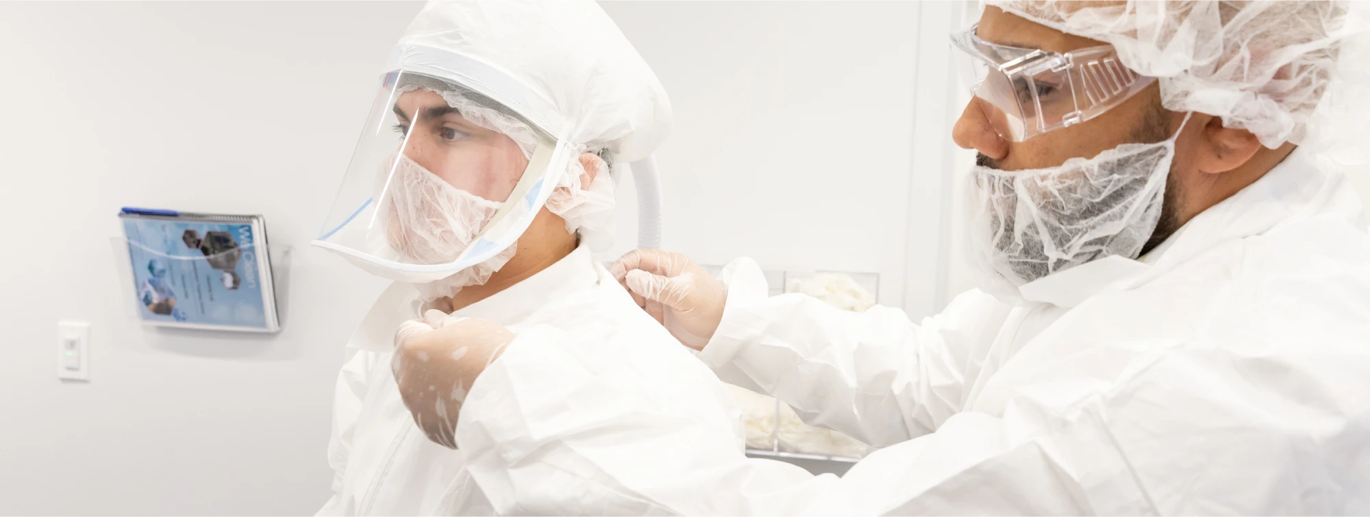 Worker in a cleanroom suit helping a colleague adjust personal protective equipment
