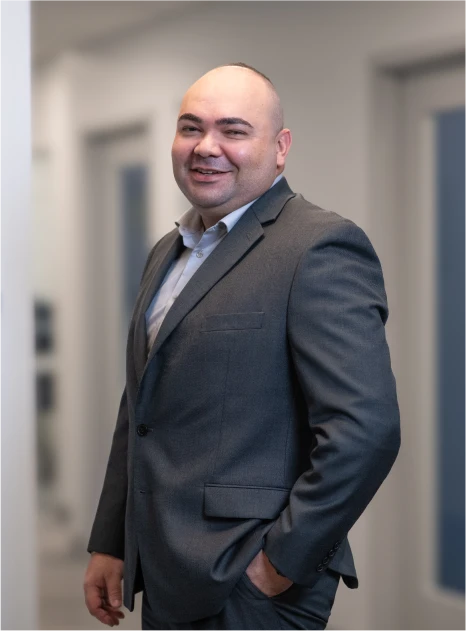 Corporate photo of man in gray jacket, open-collar shirt standing in hallway with soft lighting