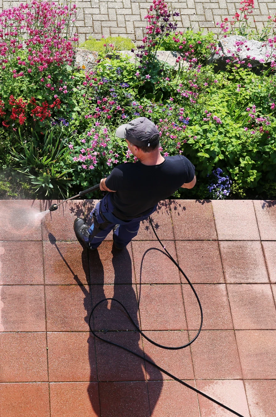 Worker using a pressure washer to clean an outdoor tiled walkway beside a lush garden