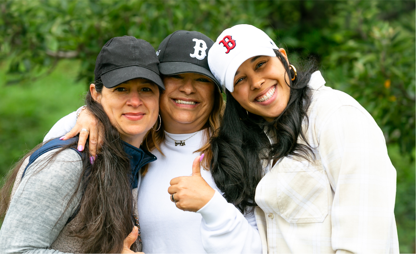 Three women in baseball caps pose outdoors, smiling and giving a thumbs-up
