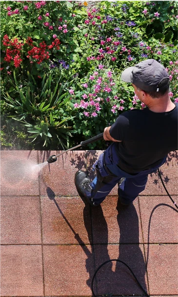Maintenance worker power-washing an outdoor pavement area surrounded by blooming flowers