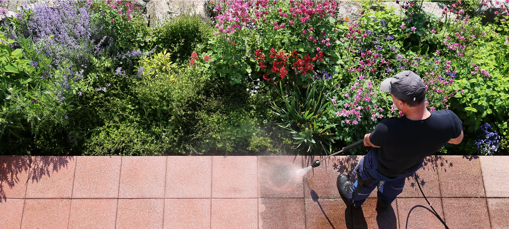 Worker using a pressure washer to clean an outdoor tiled patio near a garden